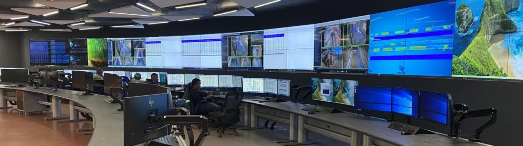 Operators at curved consoles facing a wide video wall in the Khumani mine control room